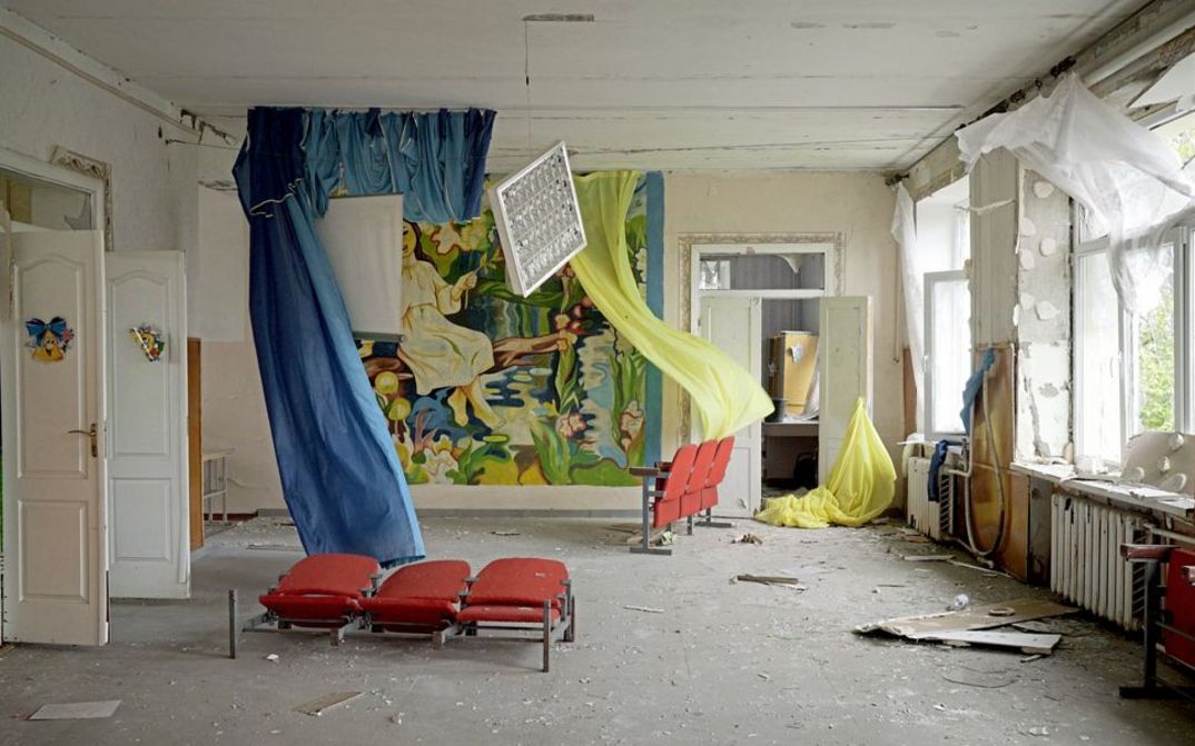 Filmstill from "Intercepted" by Oksana Karpovych. It shows an empty room with broken walls and dirt on the floor. Blue and yellow curtains hang in the room and there are red movie theater seats.
