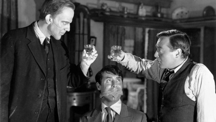 Filmstill aus ARSEN UND SPITZENHÄUBCHEN: Zwei Männer prosten einander zu, einer hat eine unheimlich aussehnde Narbe auf der Wange. Zwischen ihnen sitzt ein gefesselter und geknebelter Mann und blickt zu den beiden hoch. 