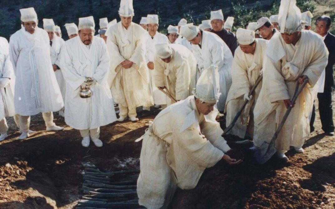 Film still from FESTIVAL: A group of men in white clothing are digging a grave.