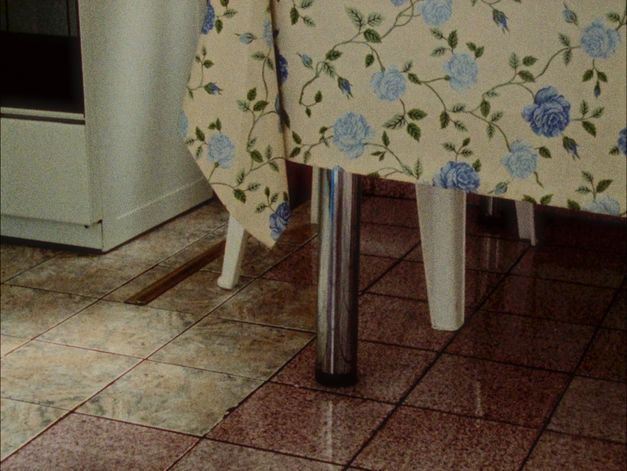 Film still from Gernot Wieland’s film “The Perfect Square”. A clean, tiled floor, metal table and plastic chair legs, a cream tablecloth with patterns of blue roses. 