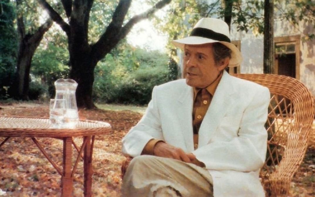 Film still from MARCELLO MASTROIANNI: MI RICORDO, SÌ, IO MI RICORDO: Marcello Mastroianni is sitting at a table in a garden in a light suit and straw hat.