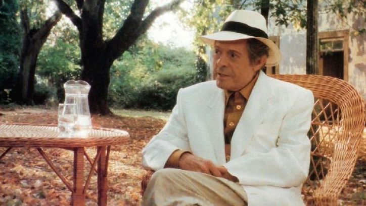 Film still from MARCELLO MASTROIANNI: MI RICORDO, SÌ, IO MI RICORDO: Marcello Mastroianni is sitting at a table in a garden in a light suit and straw hat. 