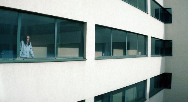 Eine Person steht hinter großen Fenstern eines modernen Gebäudes und blickt aus einem oberen Stockwerk nach außen.