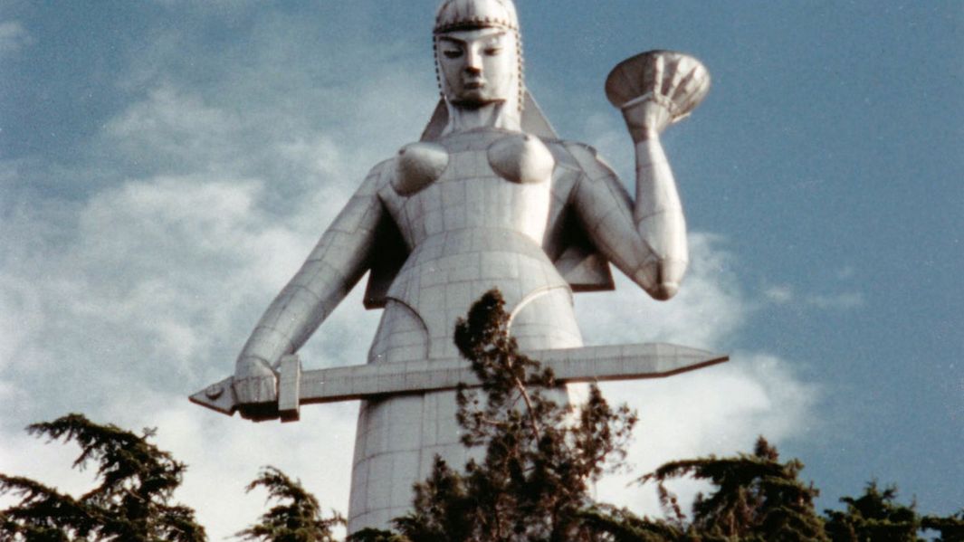 Filmstill aus DURING THE MANY YEARS: Eine riesige silbrige Statue einer Kriegerin mit einem Schwert ragt in den Himmel. Im Vordergrund sind Baumwipfel zu sehen.