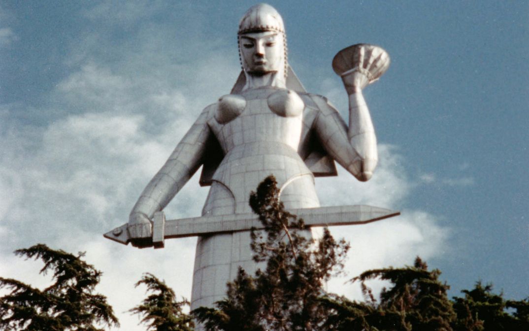Filmstill aus DURING THE MANY YEARS: Eine riesige silbrige Statue einer Kriegerin mit einem Schwert ragt in den Himmel. Im Vordergrund sind Baumwipfel zu sehen.