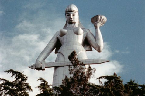 Film still from DURING THE MANY YEARS: A huge silver statue of a female warrior holding a sword towers into the sky. Tree tops can be seen in the foreground.