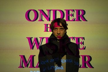 The artist Matthias De Groof stands in front of his video installation. The film title is projected onto the screen behind him and onto his body.