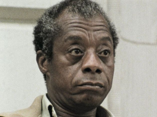 Still from the film "I Heard It through the Grapevine" by Dick Fontaine. James Baldwin, an older man with grey-brown hair and a brown jacket, is looking past the camera on the right side with an unimpressed expression.