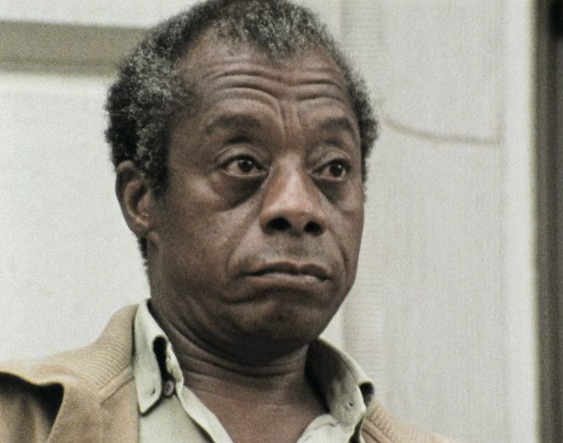 Still from the film "I Heard It through the Grapevine" by Dick Fontaine. James Baldwin, an older man with grey-brown hair and a brown jacket, is looking past the camera on the right side with an unimpressed expression.