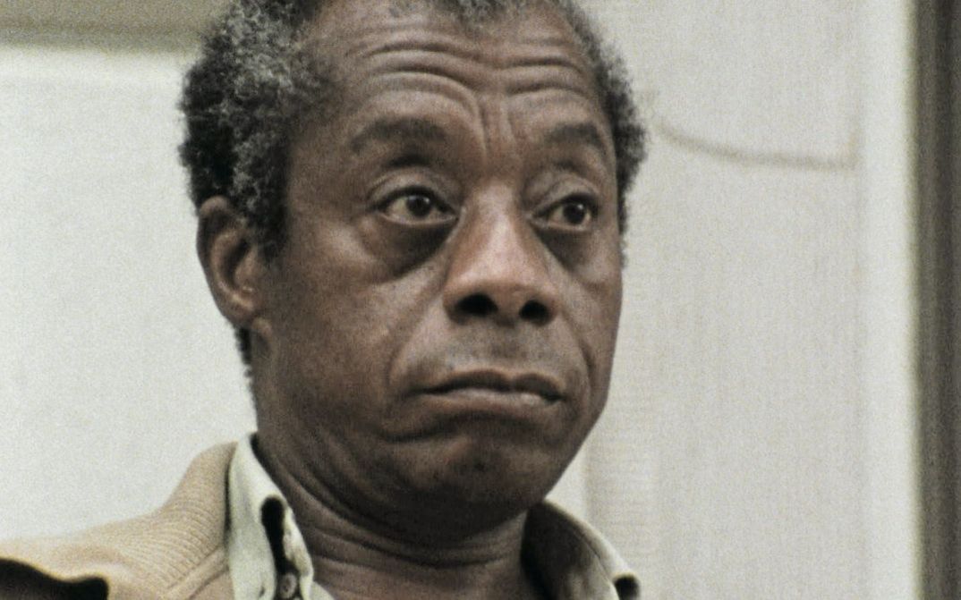 Still from the film "I Heard It through the Grapevine" by Dick Fontaine. James Baldwin, an older man with grey-brown hair and a brown jacket, is looking past the camera on the right side with an unimpressed expression.