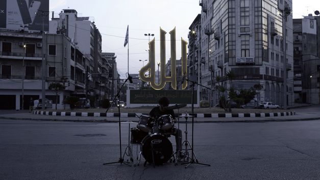 Person spielt Schlagzeug auf leerem Platz vor leuchtendem Schriftzeichen.