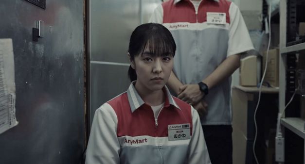 Junge Frau in Supermarktuniform sitzt in engem Lagerraum, Person steht hinter ihr.