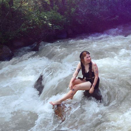 Fünf junge Frauen sitzen in Badekleidung auf Felsen in einem Fluss, rechts steigt pinker Rauch auf.