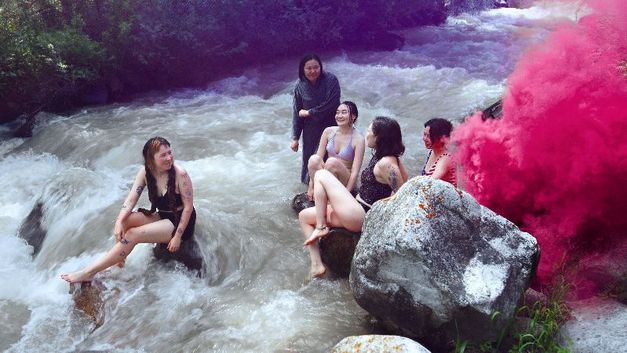 Fünf junge Frauen sitzen in Badekleidung auf Felsen in einem Fluss, rechts steigt pinker Rauch auf.
