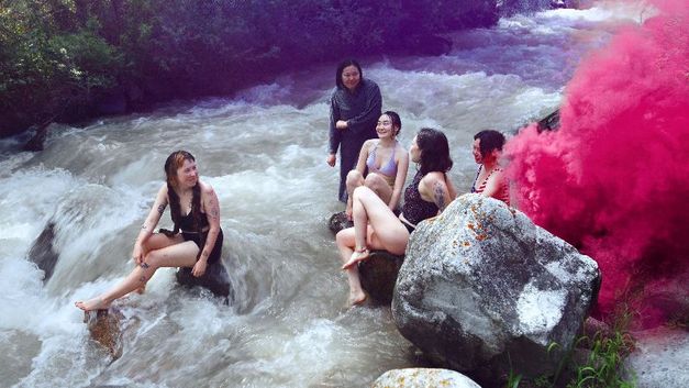 Fünf junge Frauen sitzen in Badekleidung auf Felsen in einem Fluss, rechts steigt pinker Rauch auf.