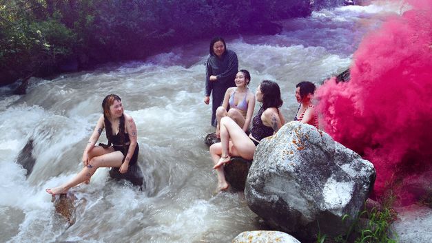 Five young women in swimwear sit on rocks in a river, pink smoke rising on the right.