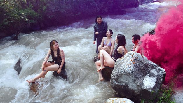Five young women in swimwear sit on rocks in a river, pink smoke rising on the right.