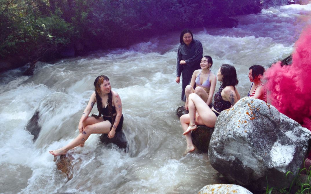 Five young women in swimwear sit on rocks in a river, pink smoke rising on the right.