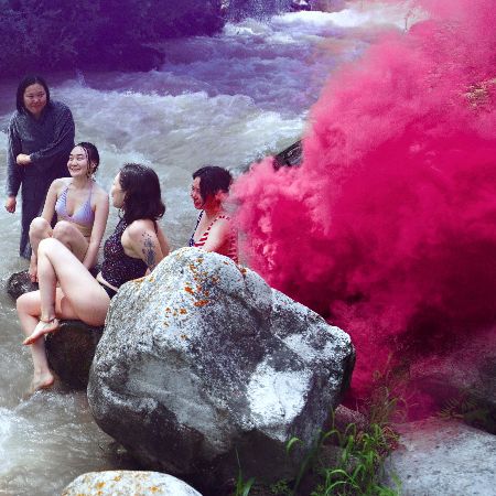 Fünf junge Frauen sitzen in Badekleidung auf Felsen in einem Fluss, rechts steigt pinker Rauch auf.
