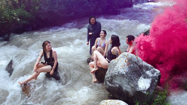 Fünf junge Frauen sitzen in Badekleidung auf Felsen in einem Fluss, rechts steigt pinker Rauch auf.