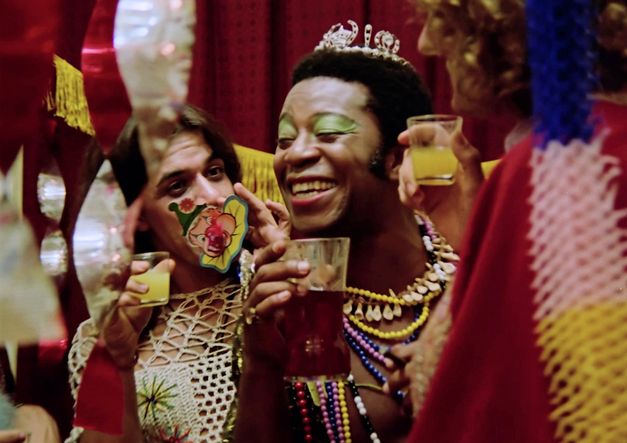 A party scene with three people wearing flamboyant costumes and saluting each other with drinks in their hands. The person in the middle is Brazil