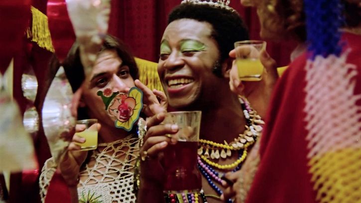 A party scene with three people wearing flamboyant costumes and saluting each other with drinks in their hands. The person in the middle is Brazil