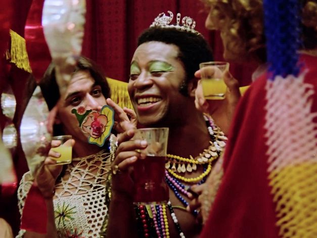 A party scene with three people wearing flamboyant costumes and saluting each other with drinks in their hands. The person in the middle is Brazil