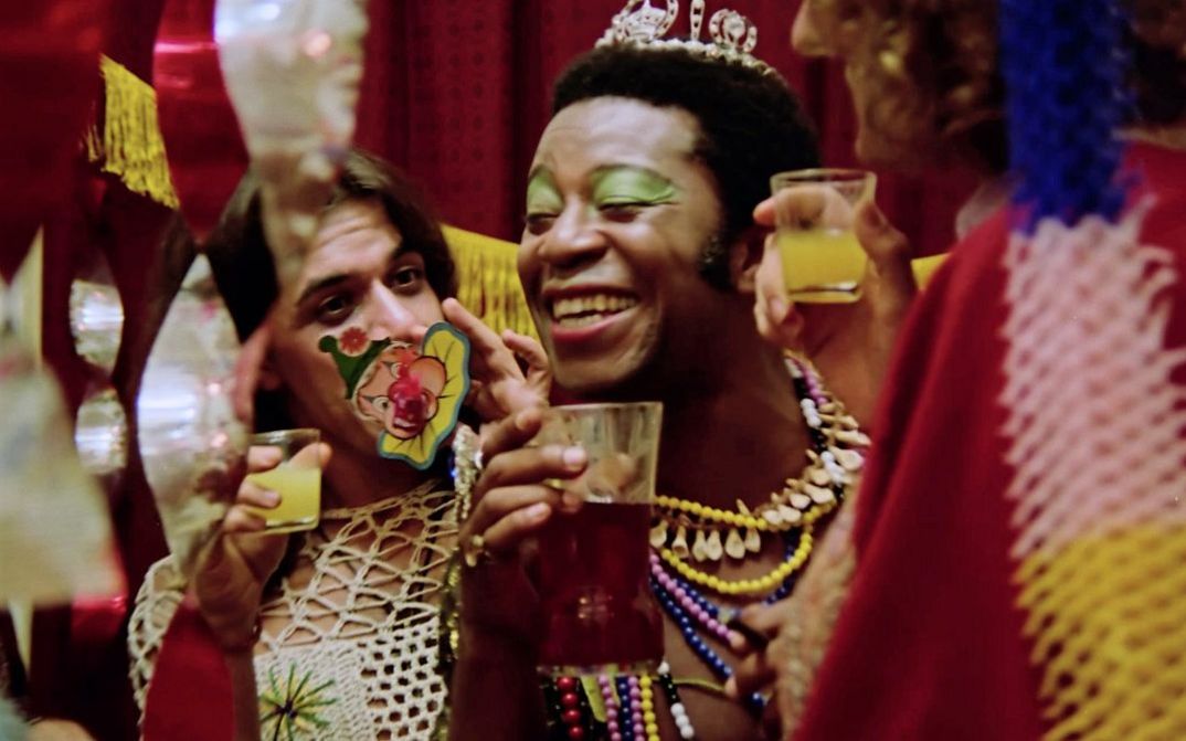 Eine Partyszene mit drei Personen, die bunte Kleidung tragen und sich mit Getränken in der Hand zuprosten. Die Person in der Mitte ist Brasiliens Schauspielstar Milton Gonçalves mit geschminktem Gesicht, einem Diadem auf dem Kopf und bunten Ketten über dem ansonsten nackten Oberkörper.