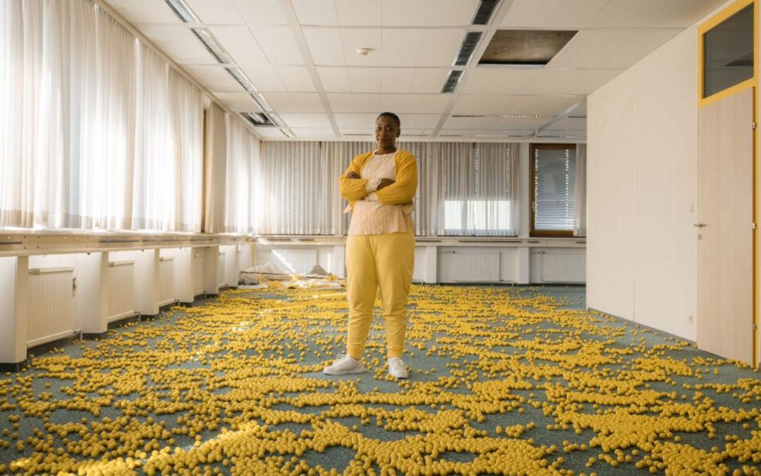 Filmstill aus FEMINISM WTF: Eine in gelb gekleidete Person steht in einem kahlen Büro voller kleiner gelber Bälle. Sie hat die Arme verschränkt und schaut in die Kamera.
