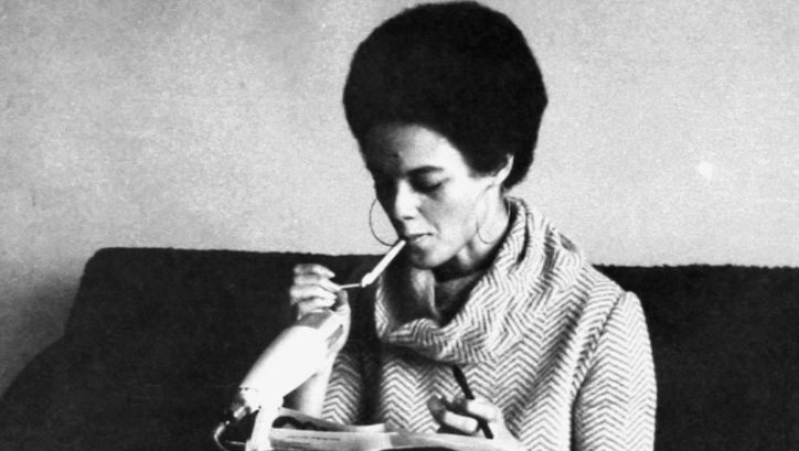 Filmstill aus KATHLEEN UND ELDRIDGE CLEAVER IN ALGIERS: Kathleen Cleavers sitzt vor einem Mikrofon und zündet sich eine Zigarette an.