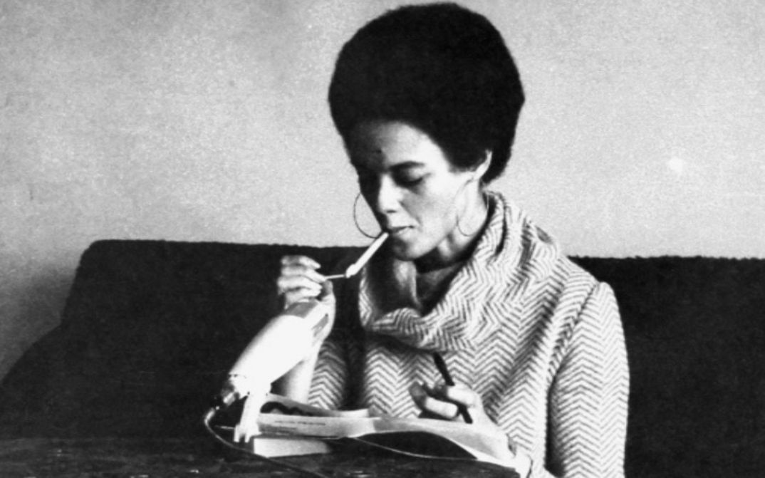 Filmstill aus KATHLEEN UND ELDRIDGE CLEAVER IN ALGIERS: Kathleen Cleavers sitzt vor einem Mikrofon und zündet sich eine Zigarette an.