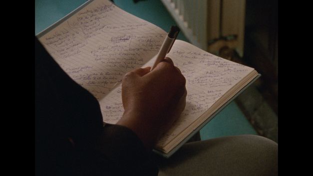 Filmstill aus dem Film „hold on to her“ von Robin Vanbesien. Eine Person schreibt diagonal auf ein liniertes Notizbuch, das mit fragmentierten Absätzen gefüllt ist.