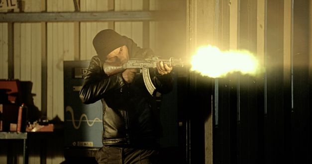 Still from the film "Le Gang des Bois du Temple" by Rabah Ameur-Zaïmeche. A man with a black hat and a black leather jacket is standing behind a wall and shooting a machine gun around the corner to the right. At the tip of the gun, there is a bright ball of light showing the explosion. 