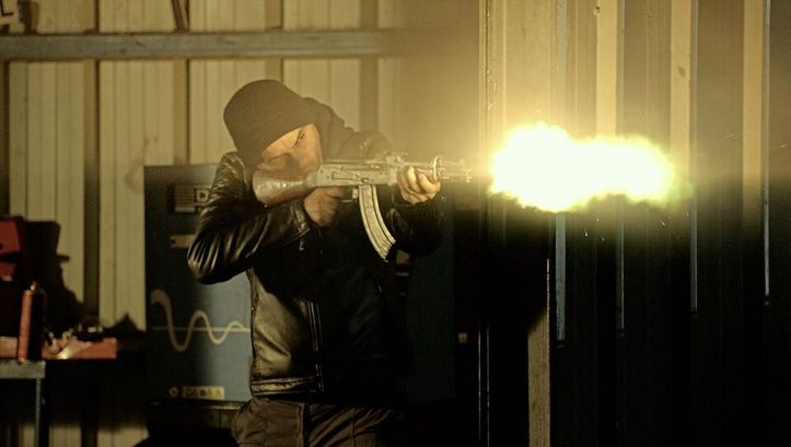 Still from the film "Le Gang des Bois du Temple" by Rabah Ameur-Zaïmeche. A man with a black hat and a black leather jacket is standing behind a wall and shooting a machine gun around the corner to the right. At the tip of the gun, there is a bright ball of light showing the explosion. 
