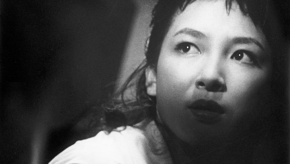 Film still from BLACK RIVER: A close-up of a young woman's face. She looks around with a frightened expression. Half of her face is hidden in shadow.