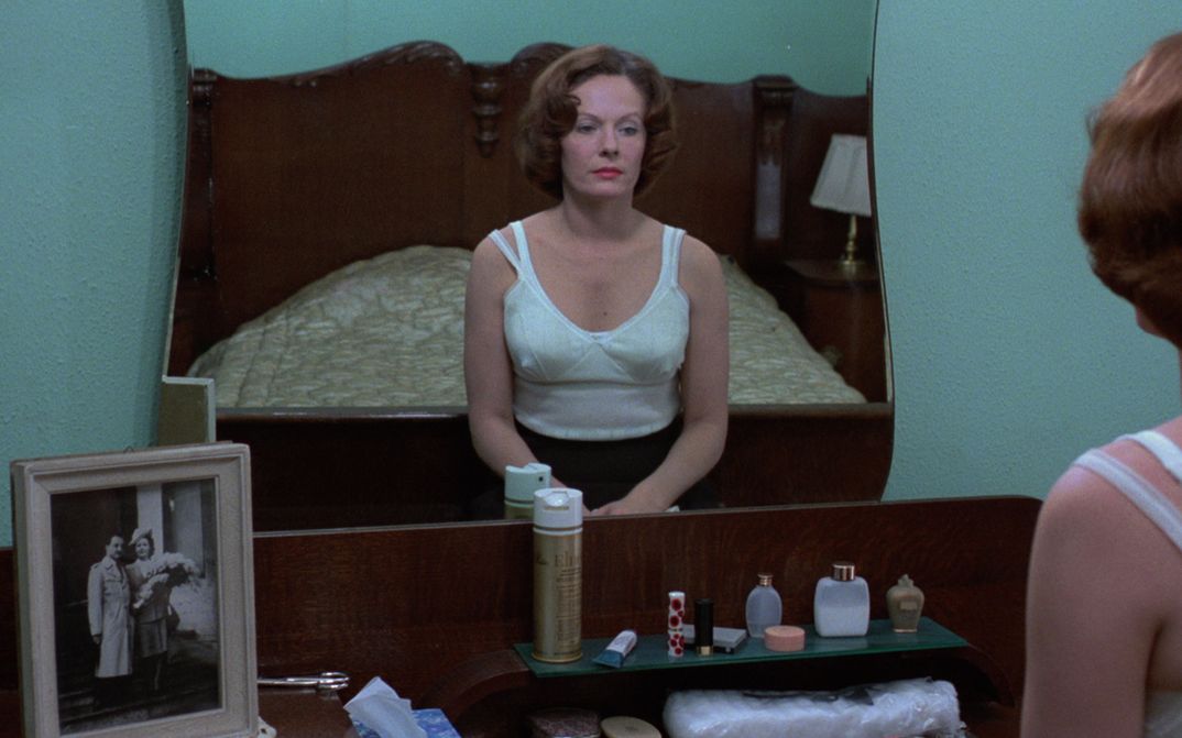 Film still from JEANNE DIELMAN, 23, QUAI DU COMMERCE, 1080 BRUXELLES: A woman sits on her bed in an undershirt and looks at herself in the mirror. 