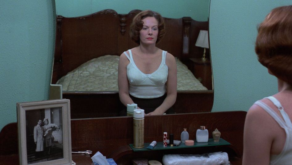 Filmstill aus JEANNE DIELMAN, 23, QUAI DU COMMERCE, 1080 BRUXELLES: Eine Frau sitzt im Unterhemd auf ihrem Bett und betrachtet sich im Spiegel.