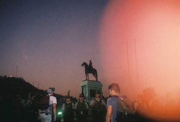 35mm colour photo at dusk, looking up at a statue of a man on a horse being protected by police. They are surrounded by young people. 