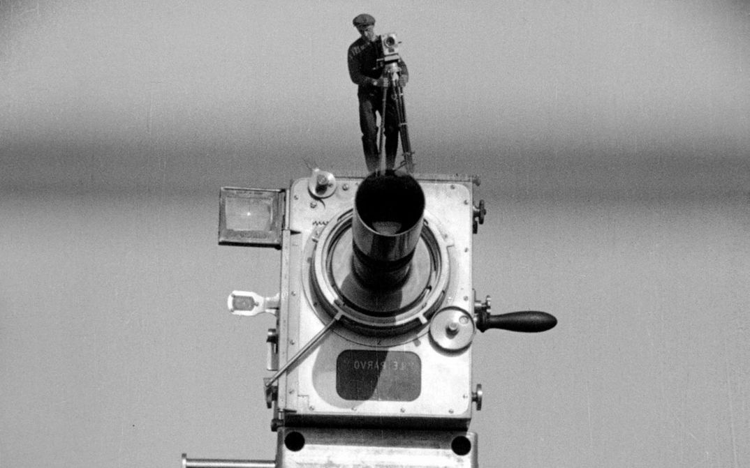 Filmstill aus MANN MIT DER KAMERA: Eine große alte Kamera, auf der ein Mann mit derselbigen Kamera steht. Er wirkt wie eine Miniatur.