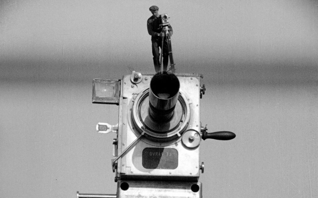 Filmstill from MANN MIT DER KAMERA: A large old camera on which a man is standing with the same camera. He looks like a miniature.