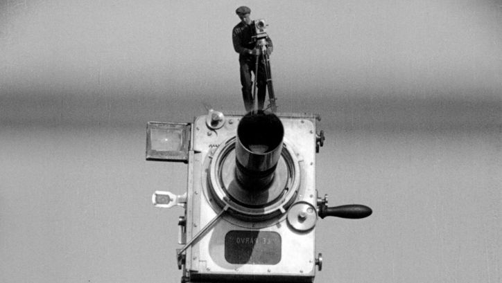 Filmstill aus MANN MIT DER KAMERA: Eine große alte Kamera, auf der ein Mann mit derselbigen Kamera steht. Er wirkt wie eine Miniatur.