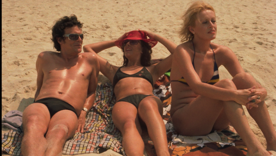 Film still from THE MAN I LOVED: wo women and a man lie on the beach in swimwear.