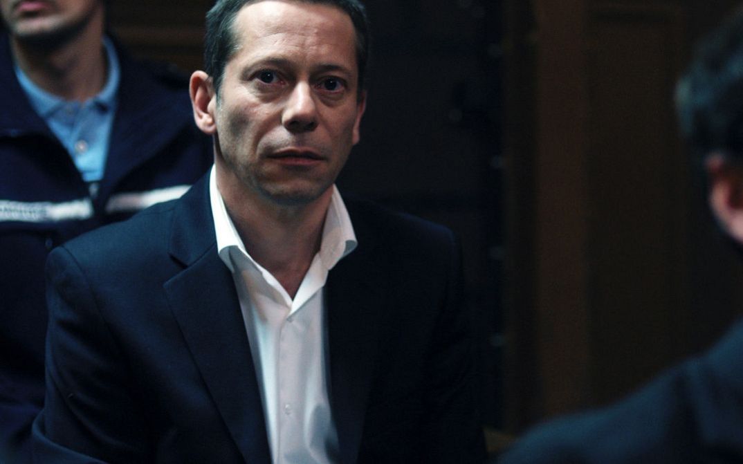 Film still from LA CHAMBRE BLEUE. A man in the courtroom, a policeman behind him.