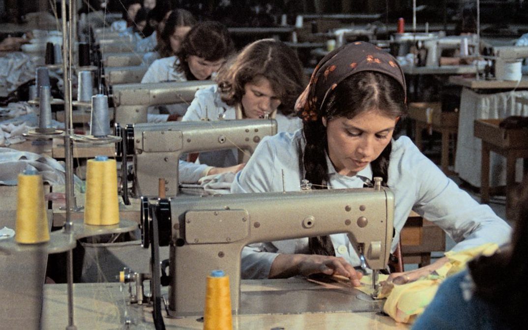 Filmstill aus „Kara Kafa“ von Korhan Yurtsever. Zu sehen ist eine Reihe von Arbeitsplätzen mit Nähmaschinen, an denen Frauen arbeiten.