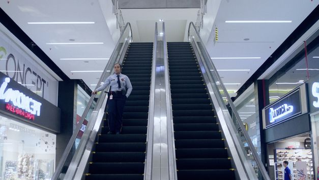 Film still from "La piel en primavera" by Yennifer Uribe Alzate. It shows two escalators in a shopping center. A woman from security is standing on the left-hand escalator, which leads downwards.