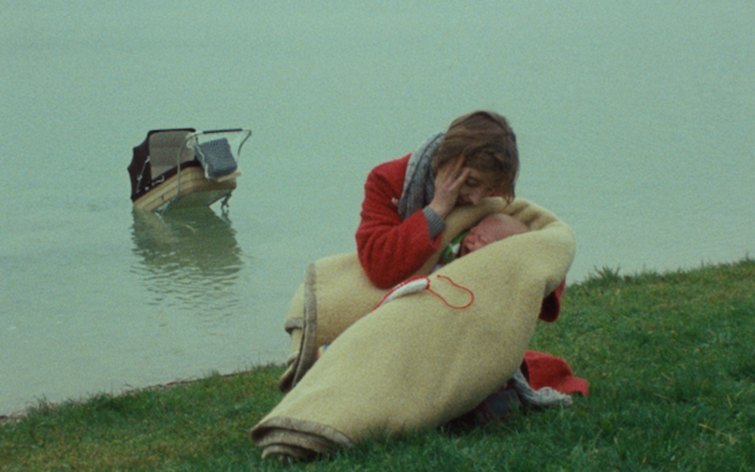 Filmstill aus EINE FRAU MIT VERANTWORTUNG: Eine Frau in einem roten Mantel hält ein schreiendes Baby in einer Wolldecke. Im Hintergrund ist ein Fluss zu sehen, in dem ein Kinderwagen schwimmt.