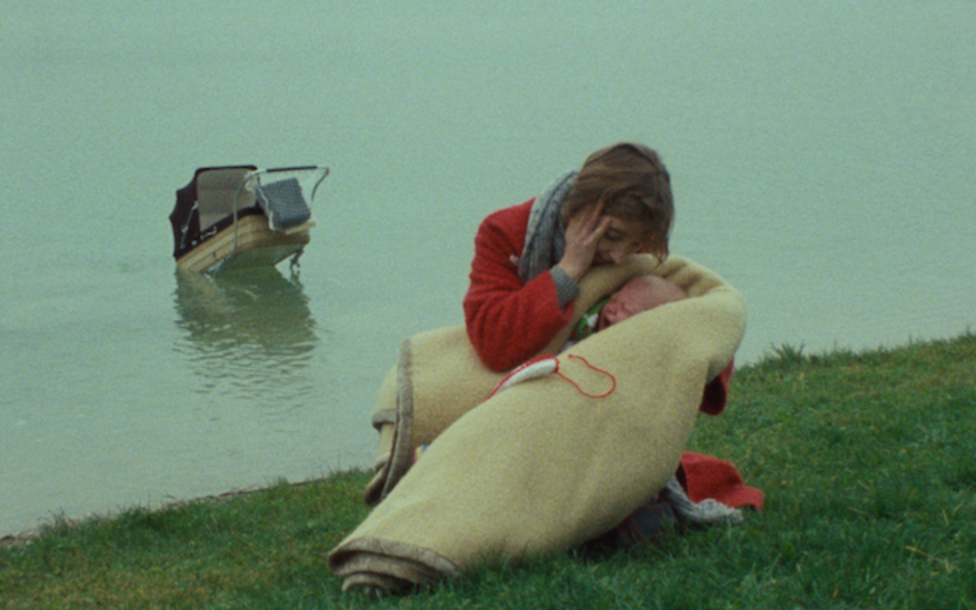 Film still from EINE FRAU MIT VERANTWORTUNG: A woman in a red coat is holding a crying baby wrapped in a wool blanket. In the background, a river can be seen with a stroller floating in it.