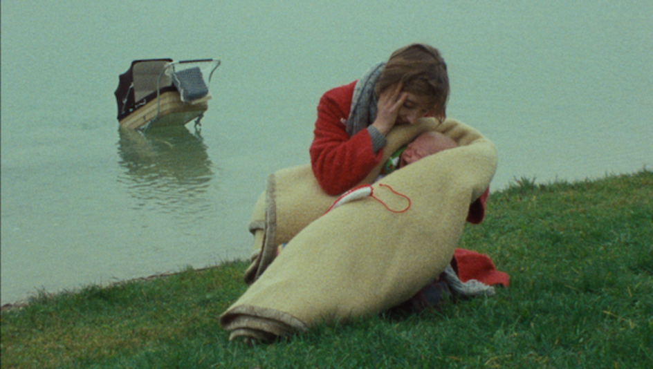 Filmstill aus EINE FRAU MIT VERANTWORTUNG: Eine Frau in einem roten Mantel hält ein schreiendes Baby in einer Wolldecke. Im Hintergrund ist ein Fluss zu sehen, in dem ein Kinderwagen schwimmt.