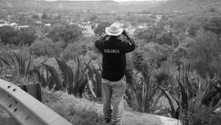 Film still from LA EMPRESA: A man looks into the desert with binoculars. He is wearing a T-shirt with the word "Vigilancia" printed on it.