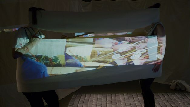 Still from the film "The Song of the Shirt" by Kerstin Schroedinger. Two people stretch a fabric between their bodies onto which images are projected.